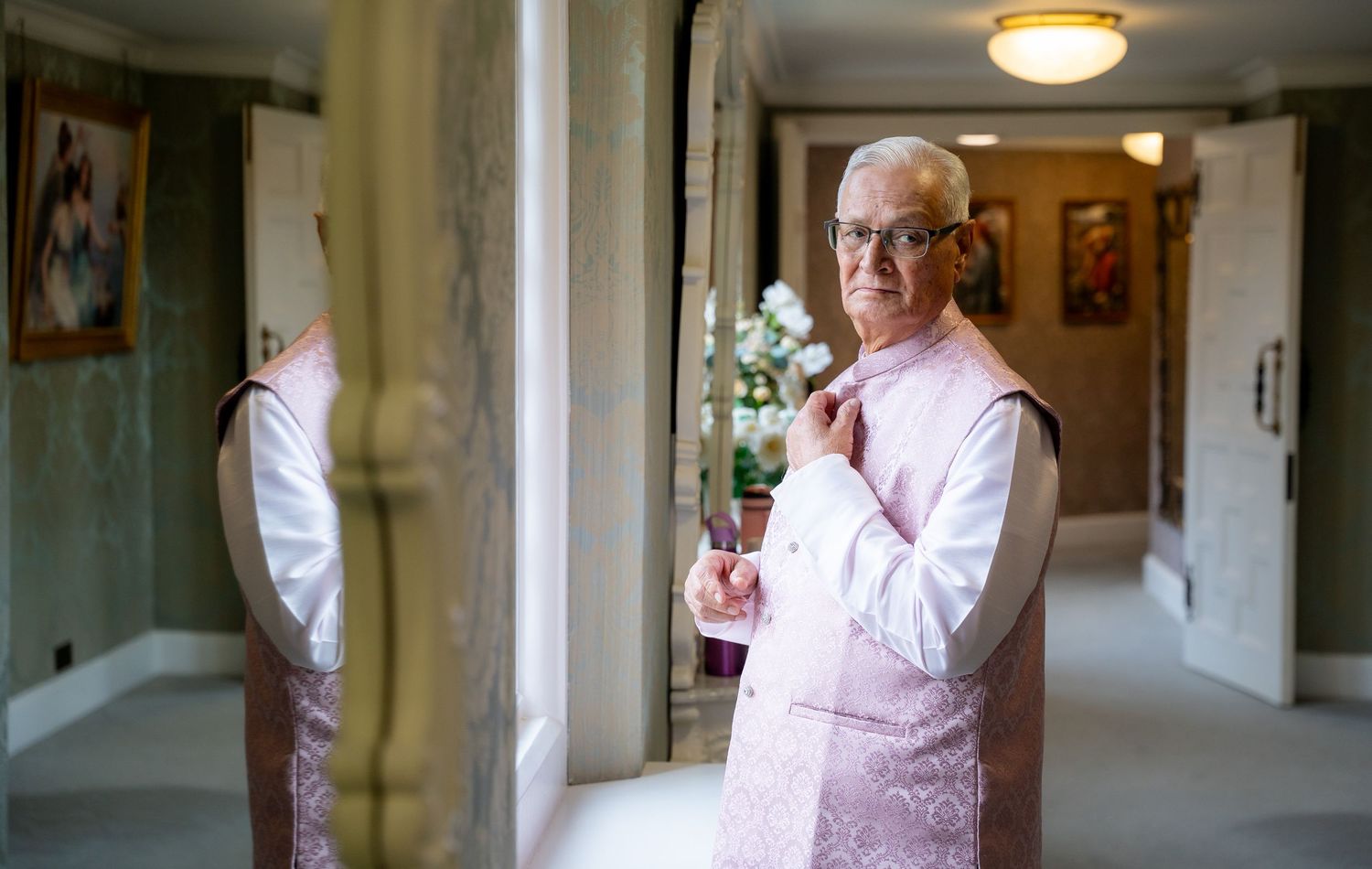 Elderly man in pink kurta adjusting tie, reflected in mirror in elegant hallway during Indian wedding.