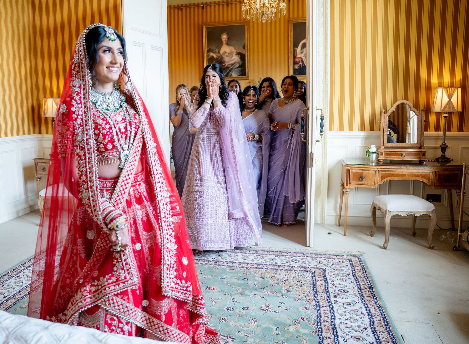 Indian bride in red lehenga reveals herself to bridesmaids in lavender dresses in grand room.