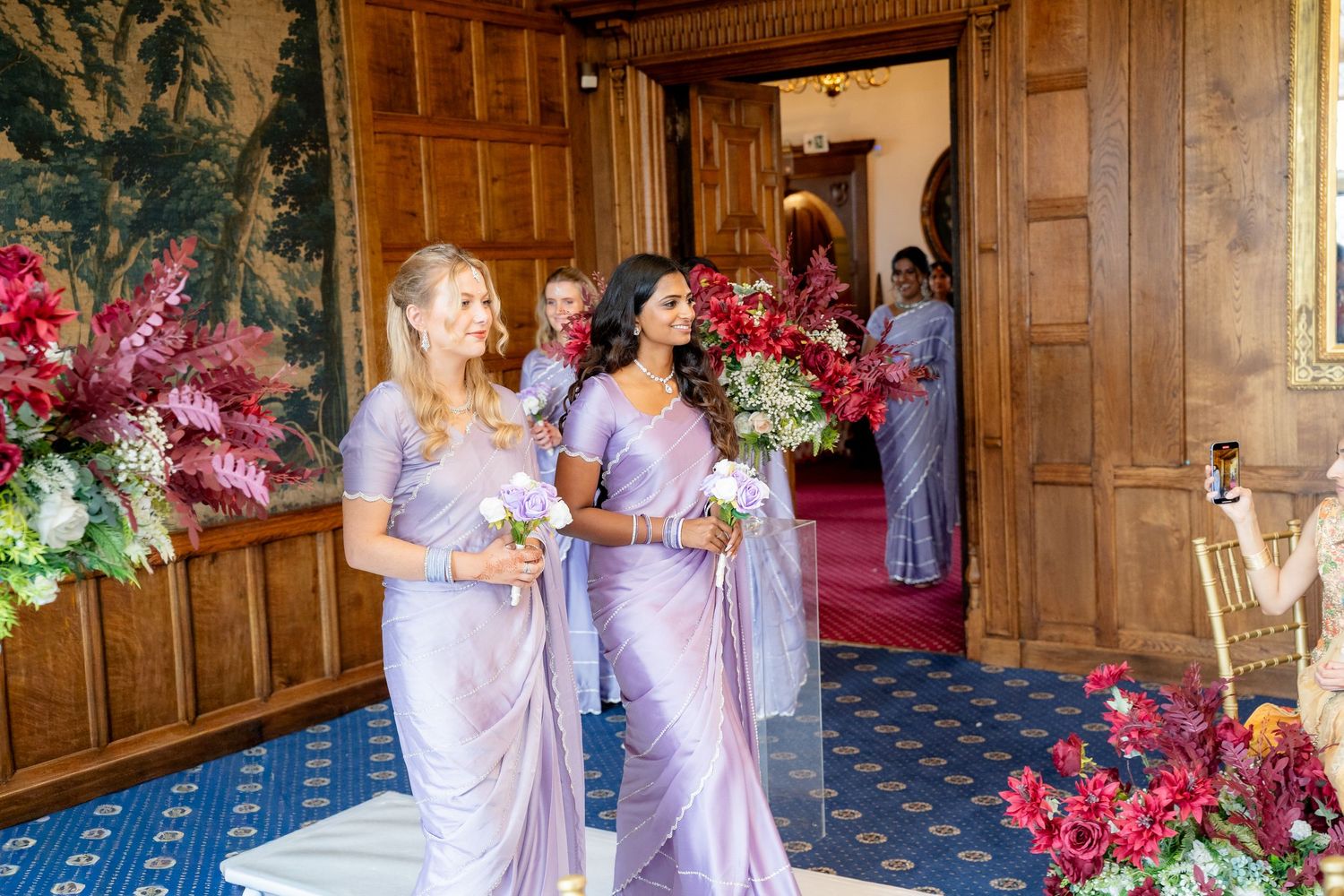 Bridesmaids in lavender sarees walk through decorated hall carrying bouquets at Indian wedding.