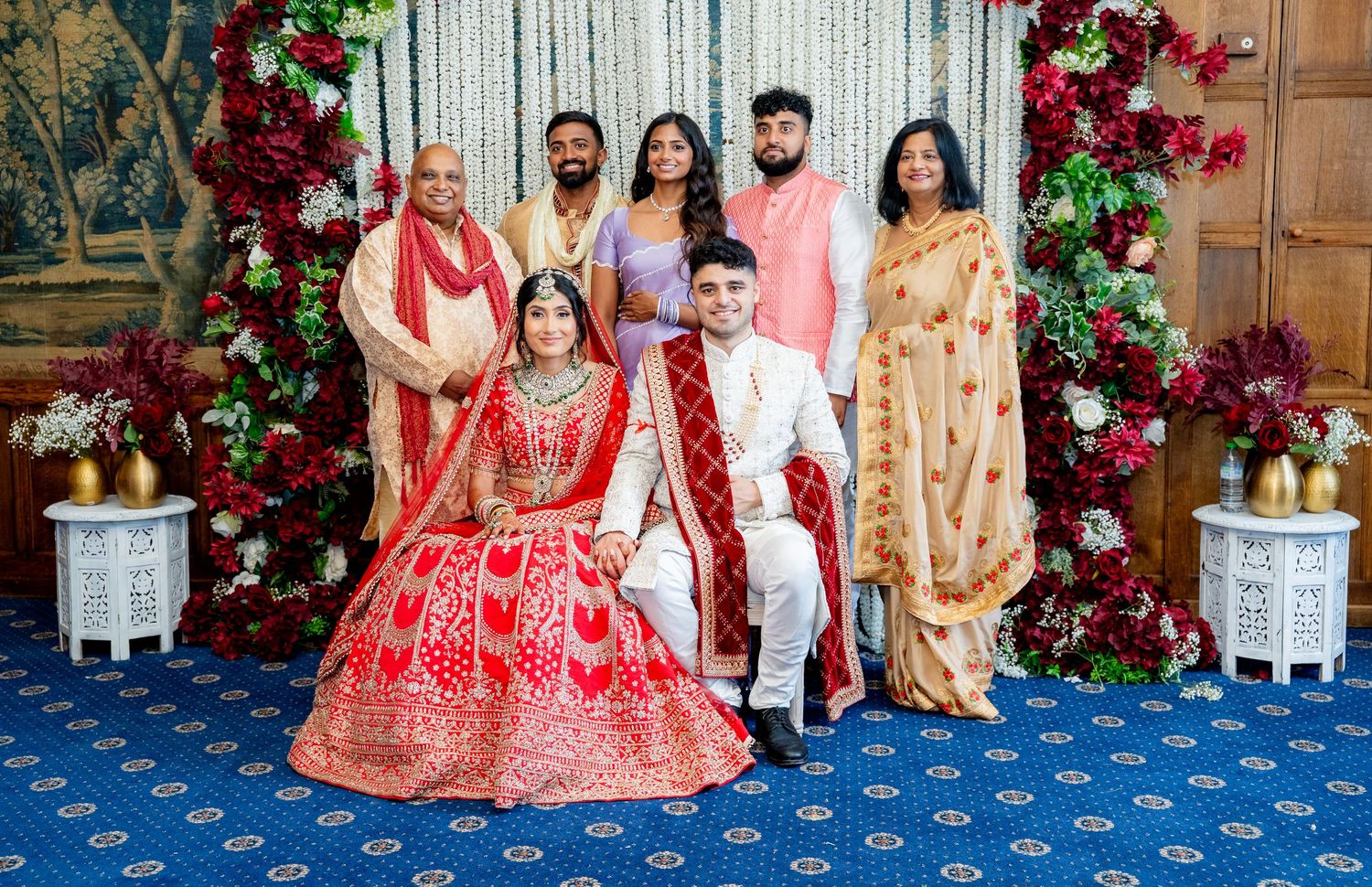 Indian bride and groom in traditional red and white attire pose with family under floral arch.