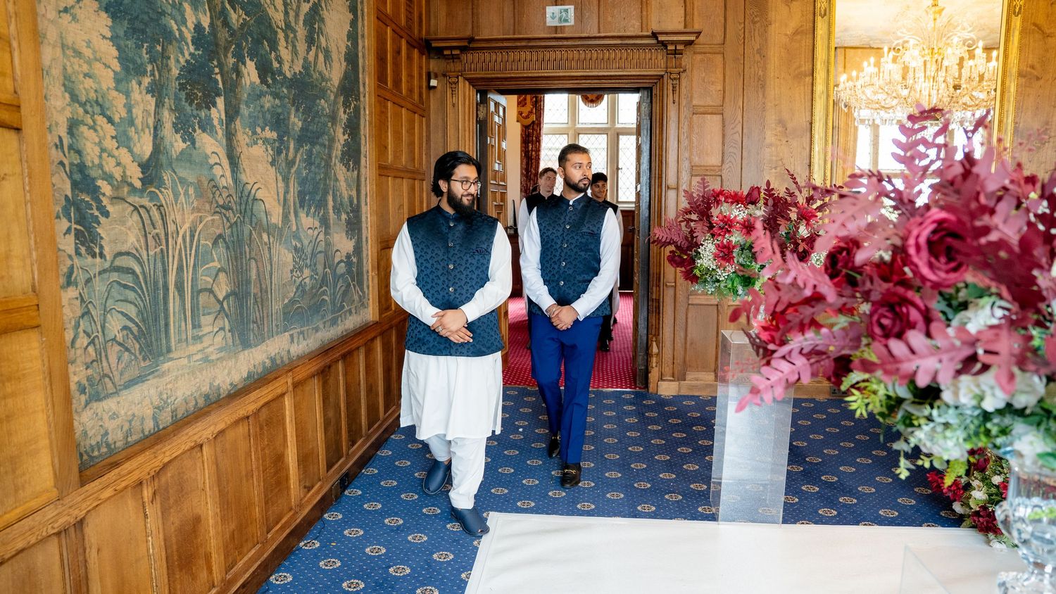 Two men in traditional Nehru jackets walk through elegant wood-paneled hall at Indian wedding.