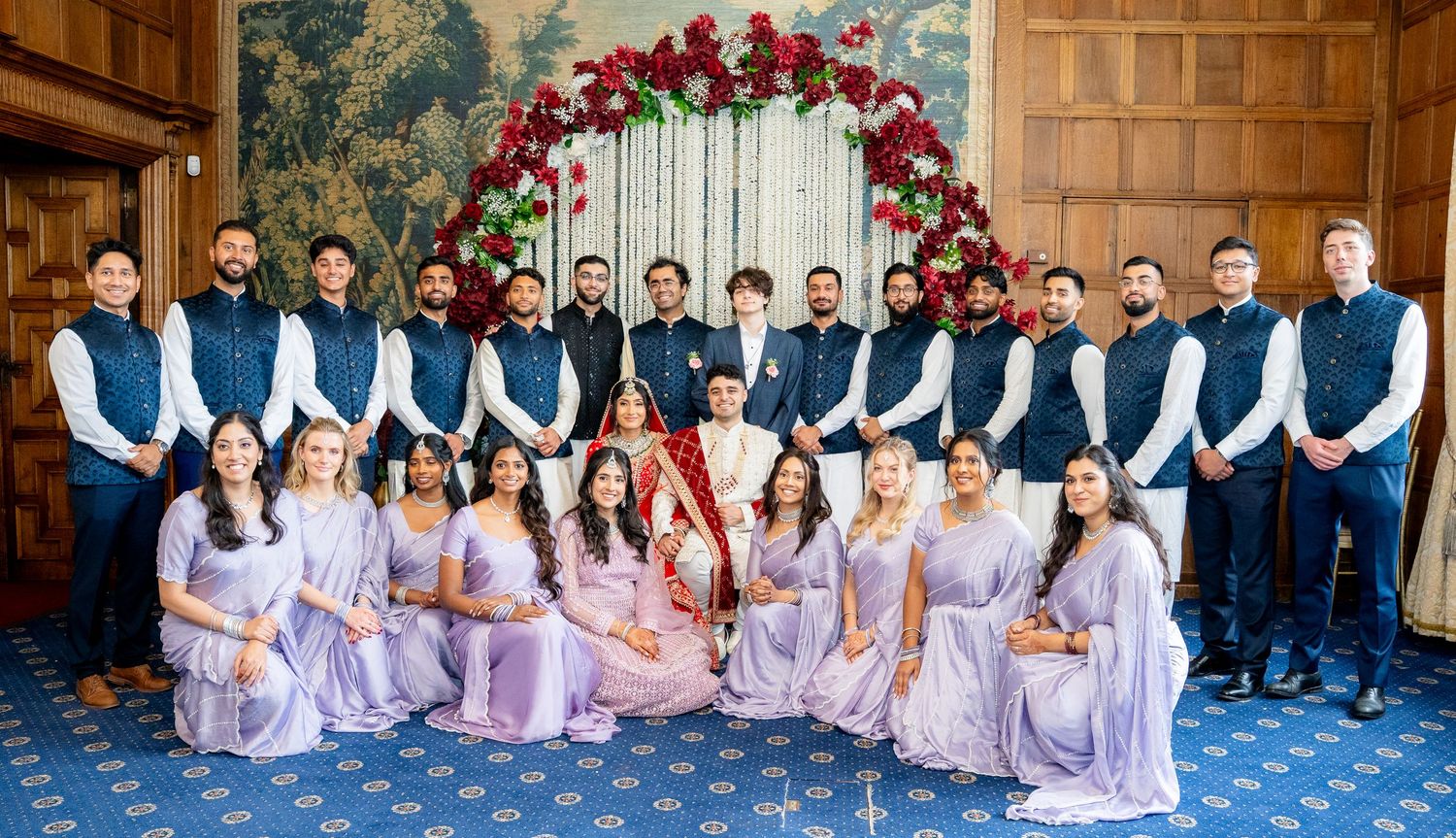 Large wedding party with bridesmaids in lavender sarees and groomsmen in navy vests posing together.