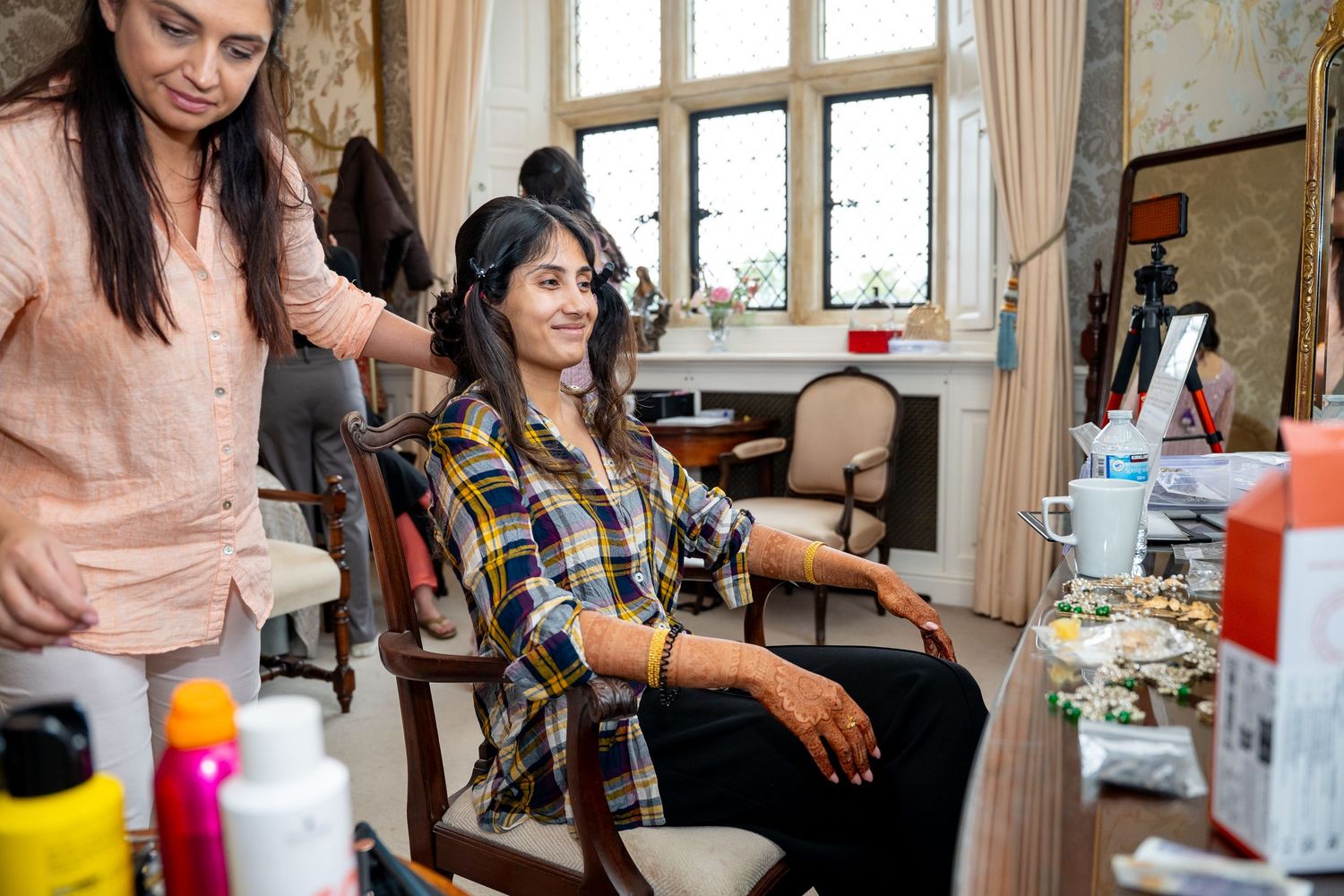 Woman getting hair styled by a beautician in a bright room with makeup products on the table.