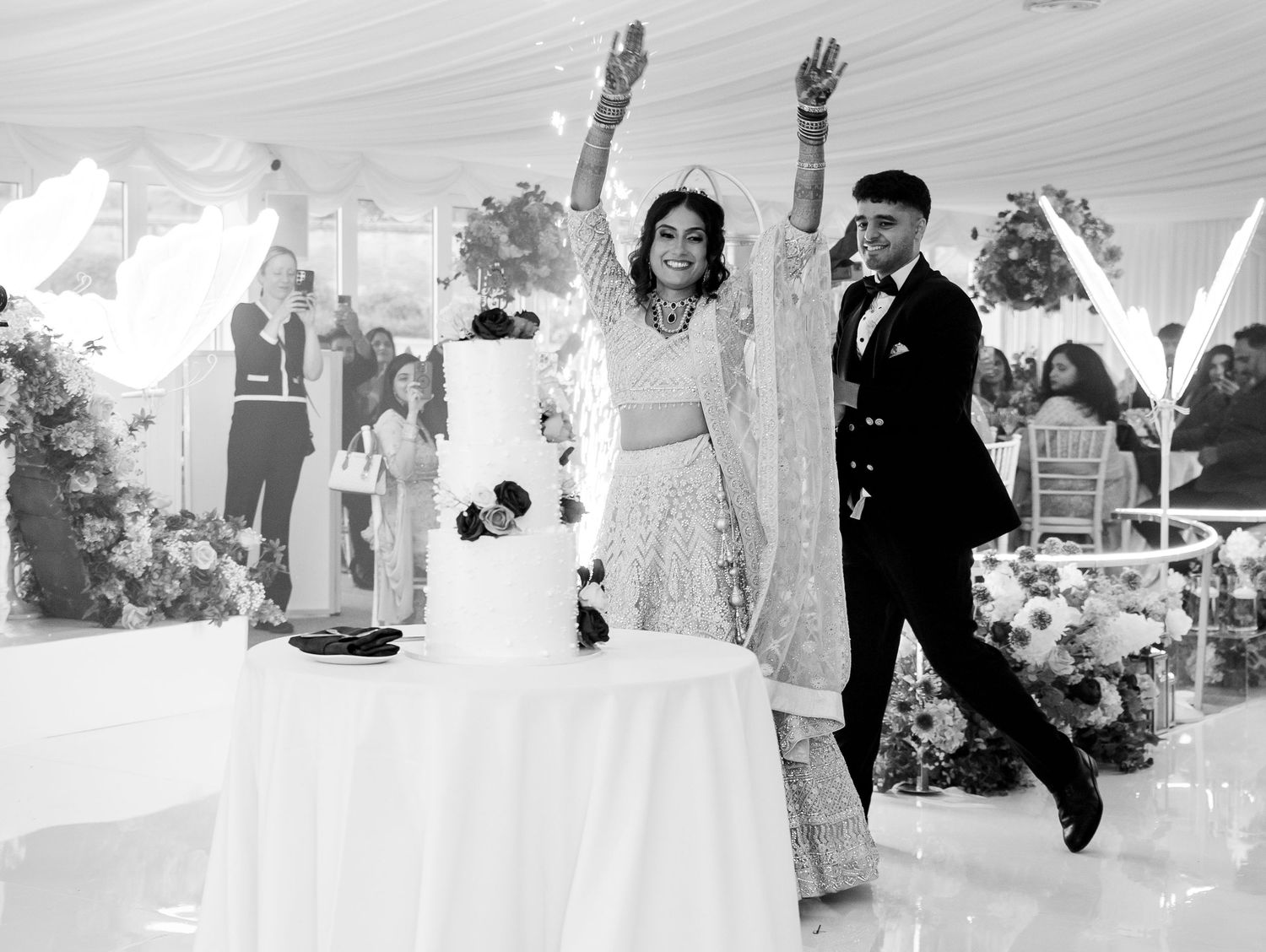 Bride and groom celebrate joyfully beside tiered wedding cake under elegant tent with floral decor in black and white photo.