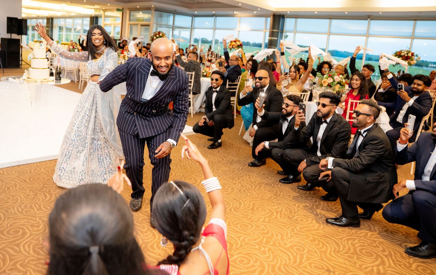 Groom in pinstripe suit reaches out to guest while dancing with bride at lively wedding reception.