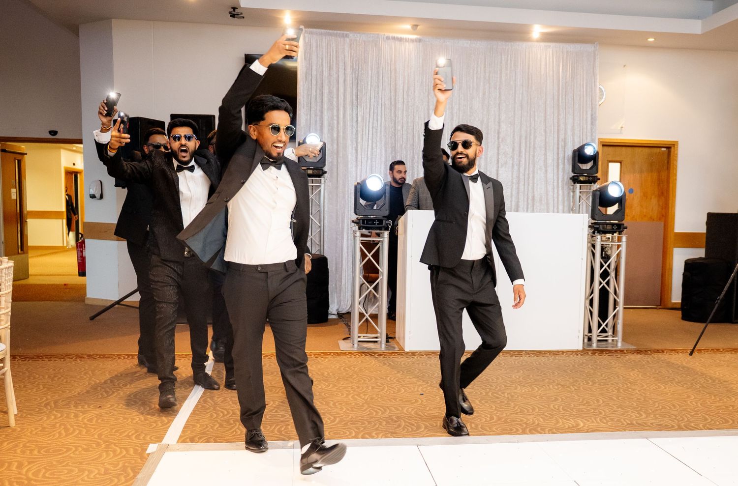 Three groomsmen in black tuxedos dancing energetically on a white dance floor at a wedding reception.