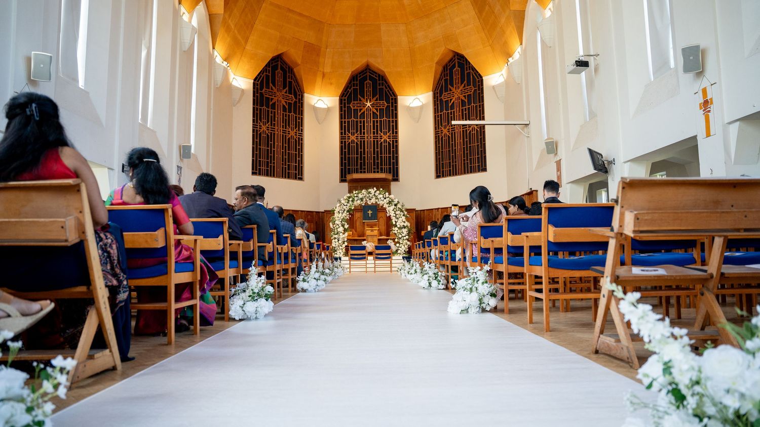 Elegant church wedding ceremony with floral-lined aisle, wooden pews, and ornate stained glass windows in the background.