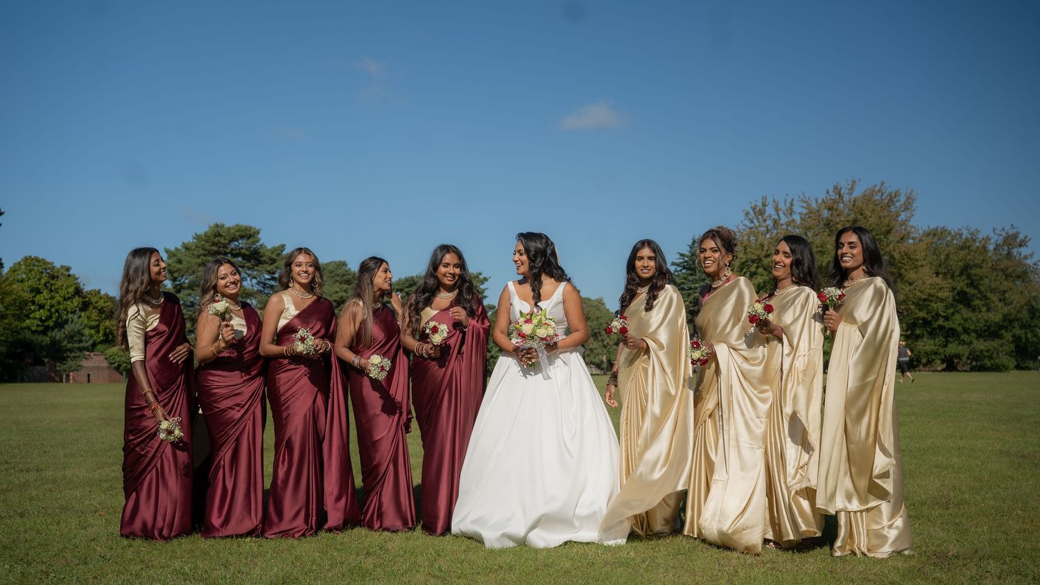 Bride surrounded by smiling bridesmaids in burgundy gowns and gold sarees on green lawn.
