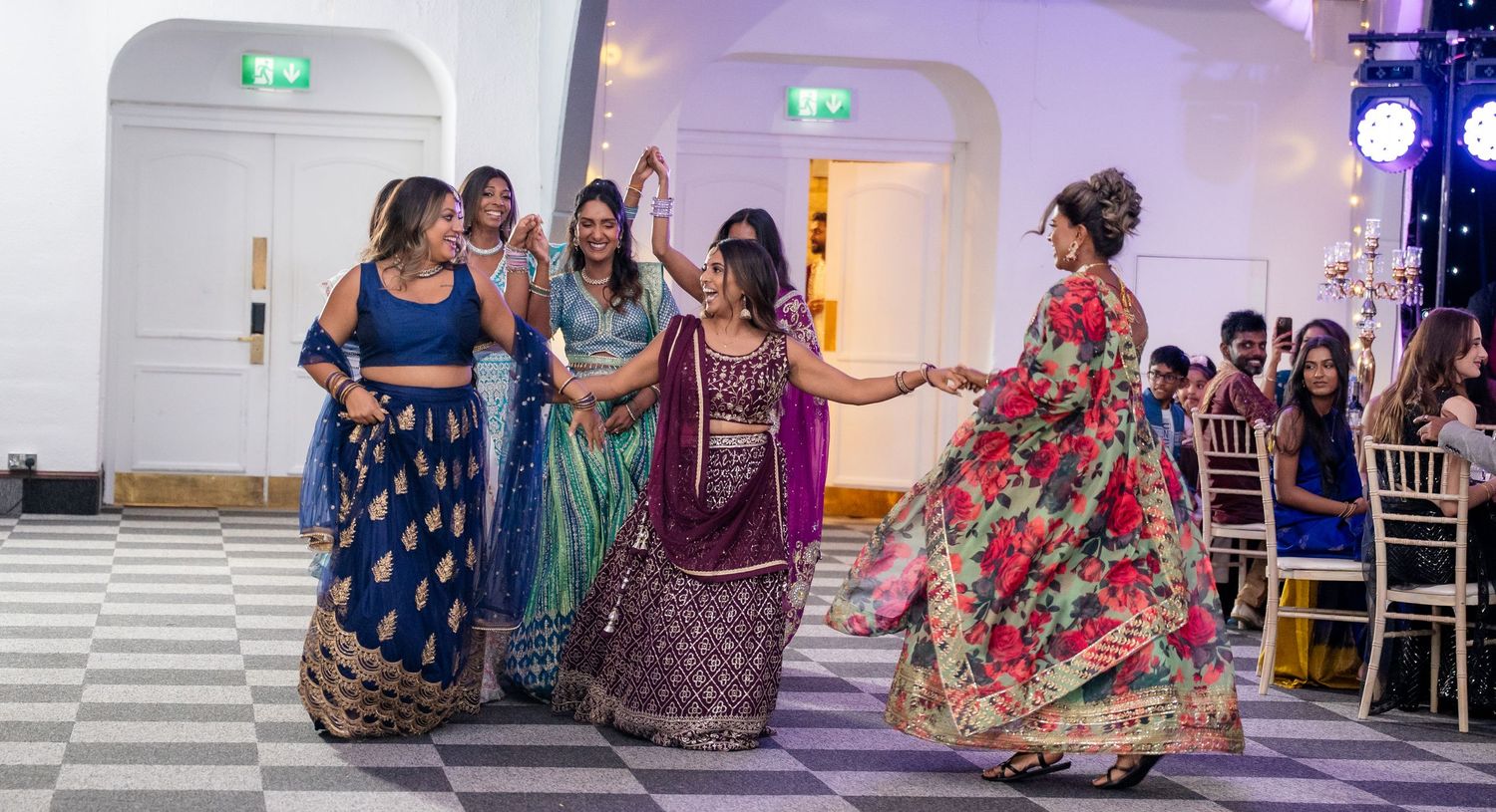 Women in colorful Indian lehengas and sarees performing a dance at a wedding celebration.