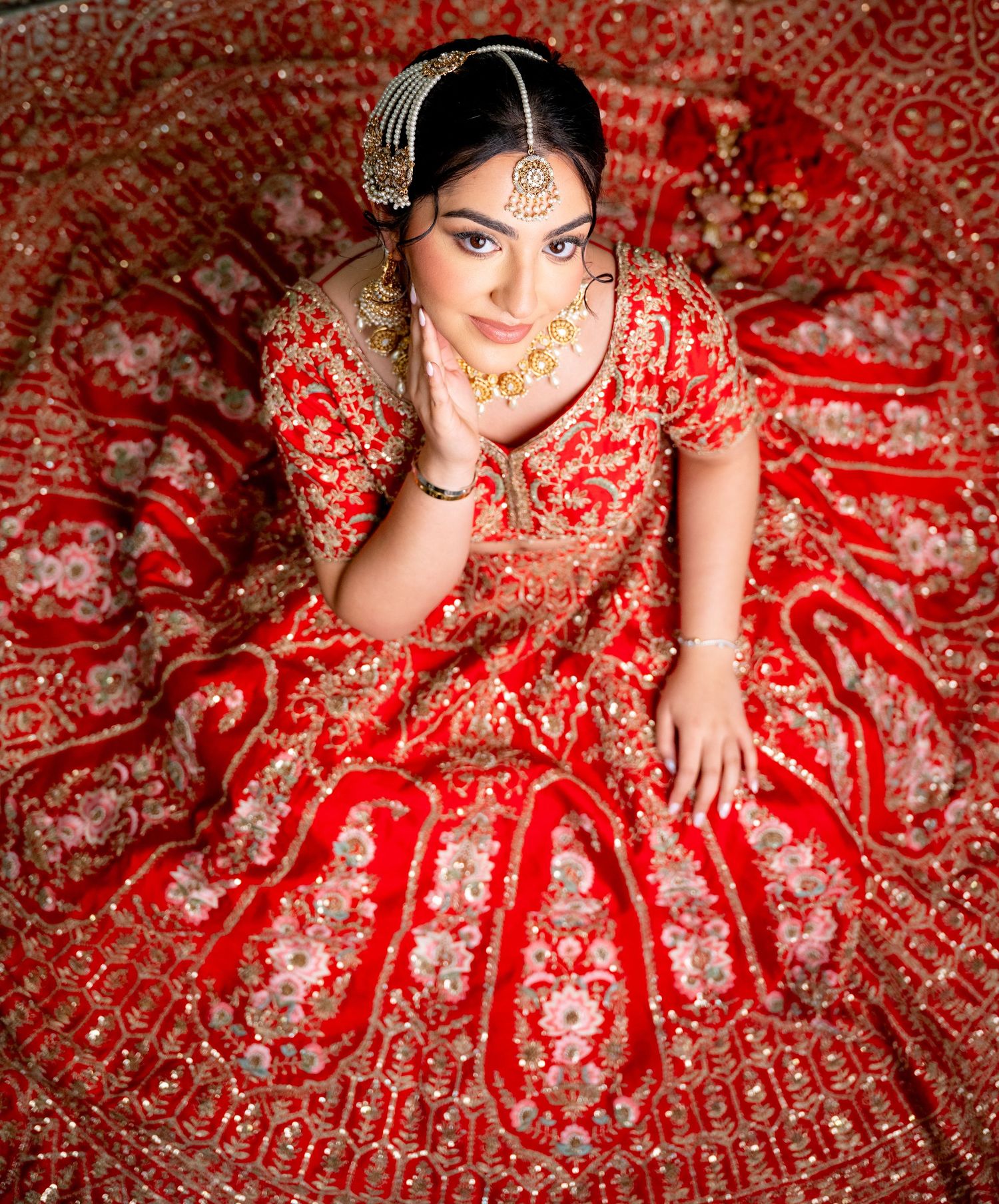 Indian bride in ornate red lehenga sits surrounded by flowing skirt with gold embroidery details.
