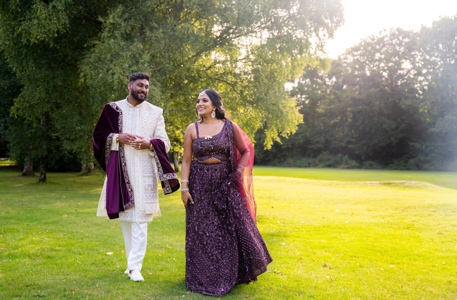 Indian wedding couple laughing and walking on lush green lawn in coordinating purple and white traditional outfits.