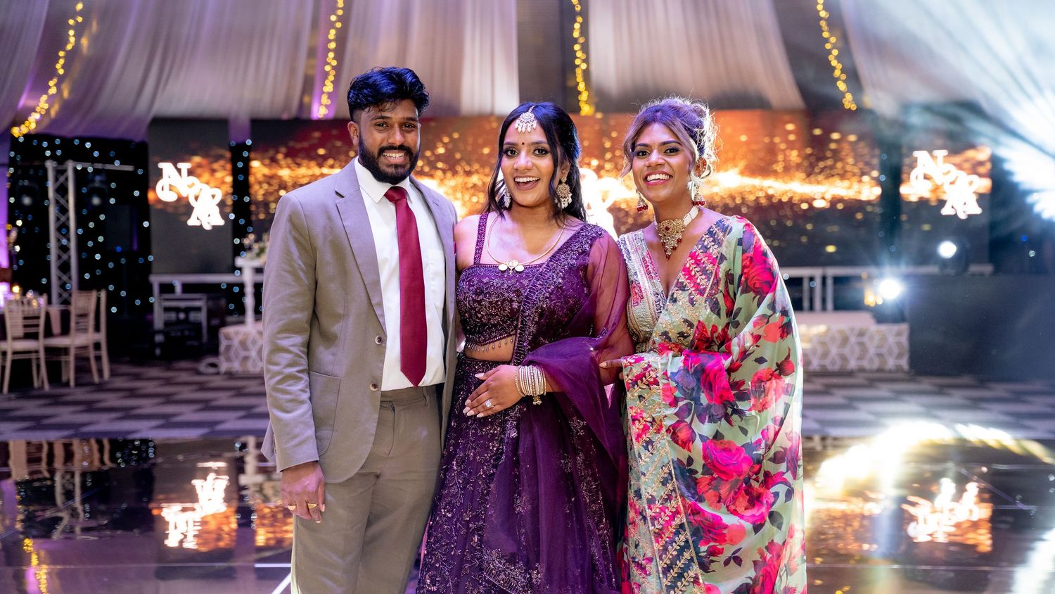 Three people posing elegantly at a festive Indian wedding reception with warm fairy lights and floral decorations.