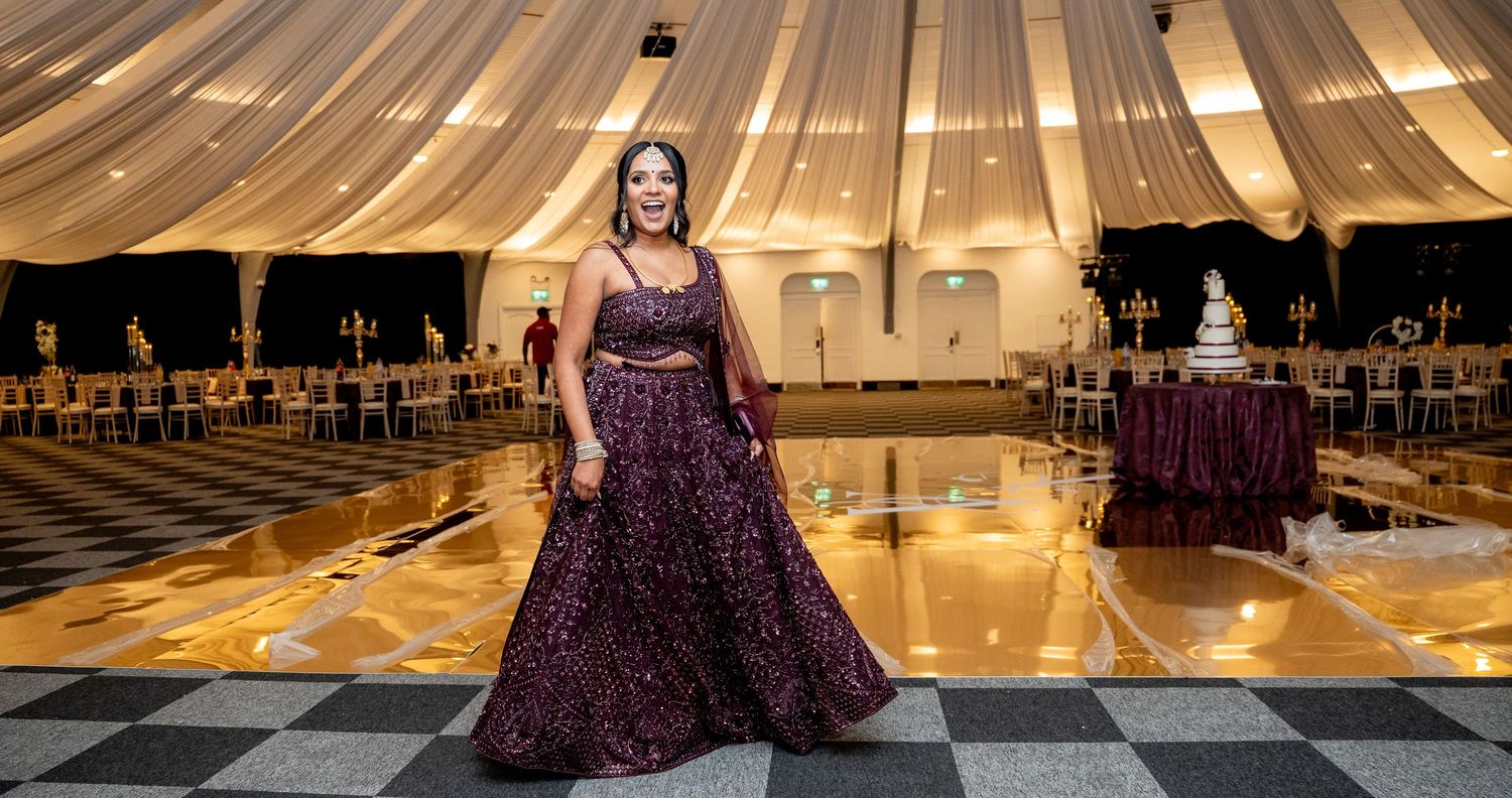 Woman in elegant purple gown poses in grand ballroom with checkered floor and draped ceiling.