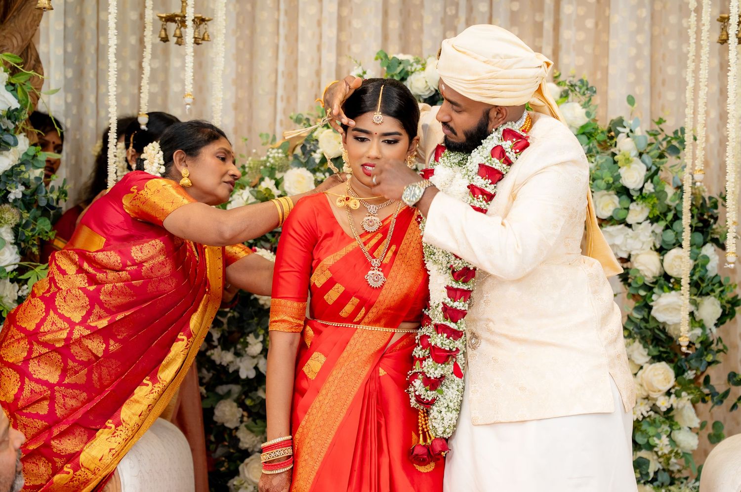 South Indian bride in orange saree receives blessings as groom in white sherwani places floral garland during wedding.