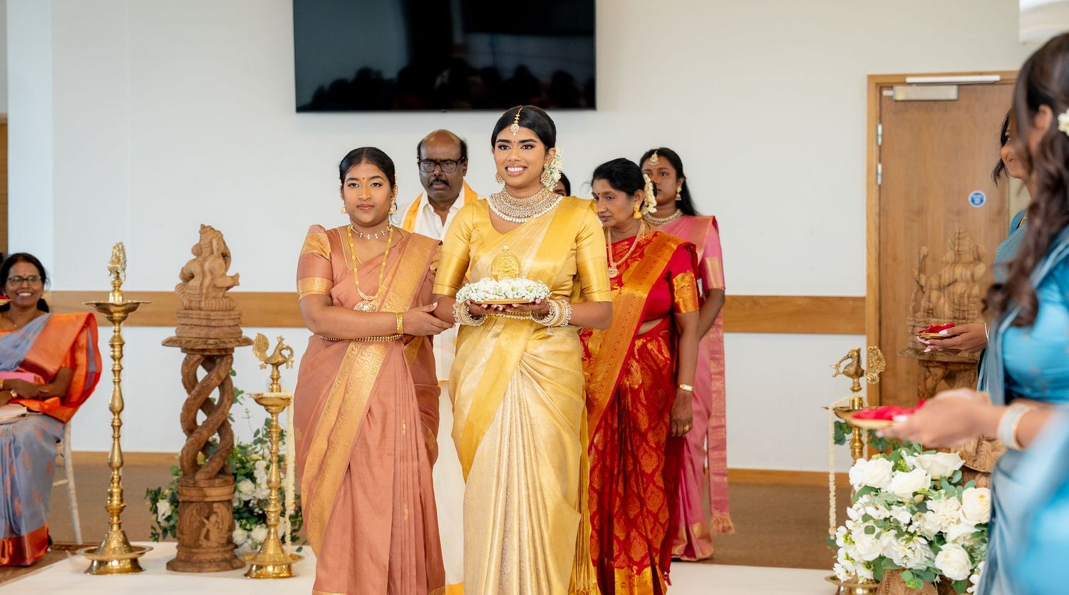 Bride in gold saree walks with family at South Indian wedding ceremony with traditional brass lamps.