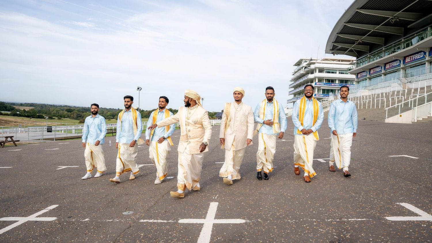Indian groom and groomsmen in traditional attire walking confidently at a racetrack venue on a cloudy day.