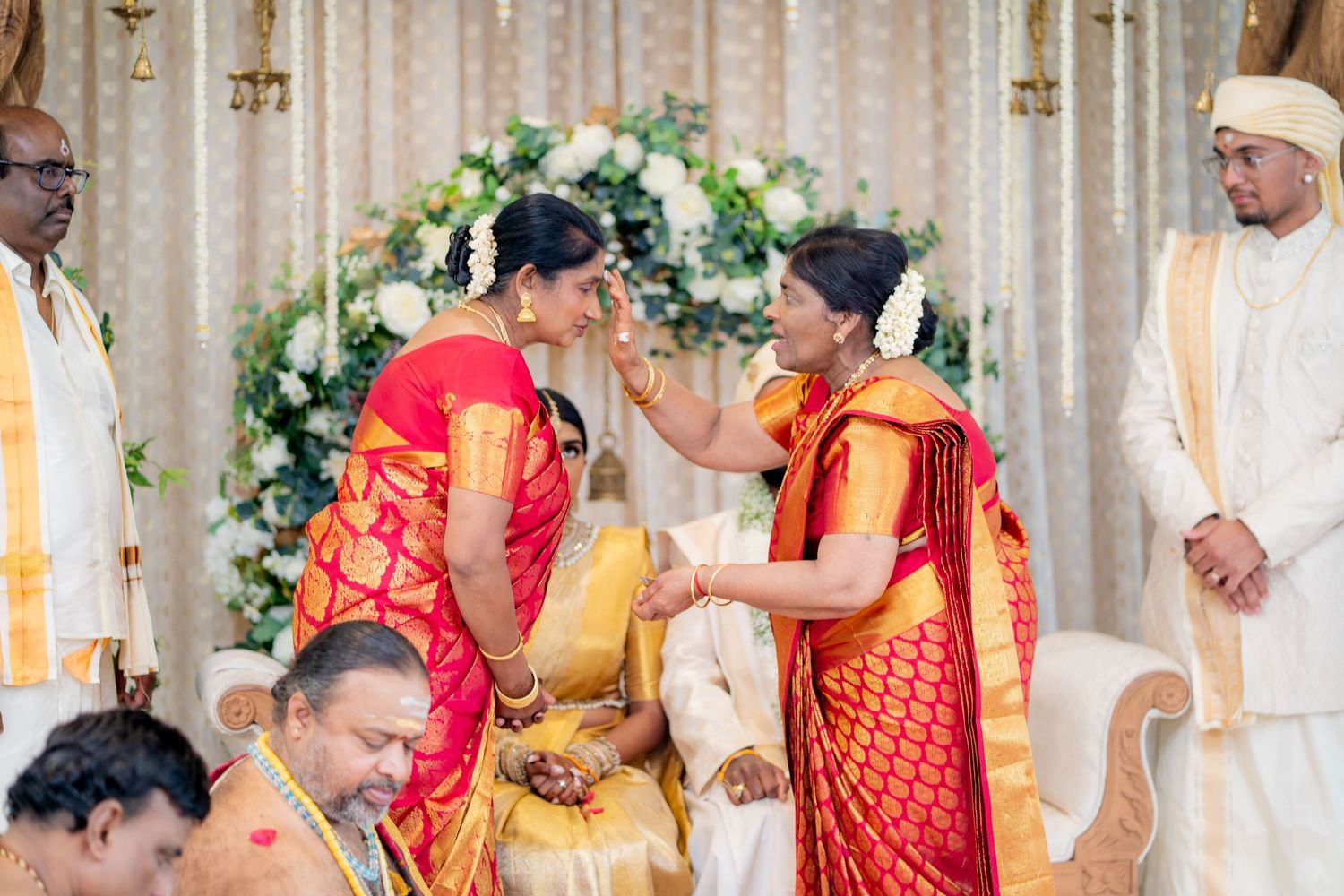 Two women in vibrant red and gold sarees share a tender moment during a traditional South Indian wedding ceremony.