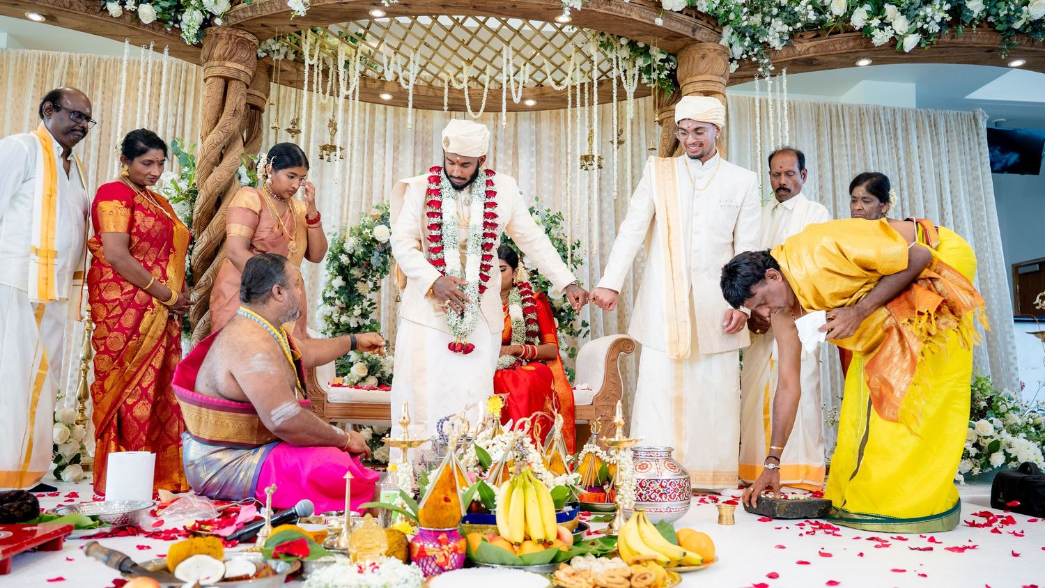 South Indian Hindu wedding ritual with priest performing puja as bride and groom stand under floral mandap decoration.