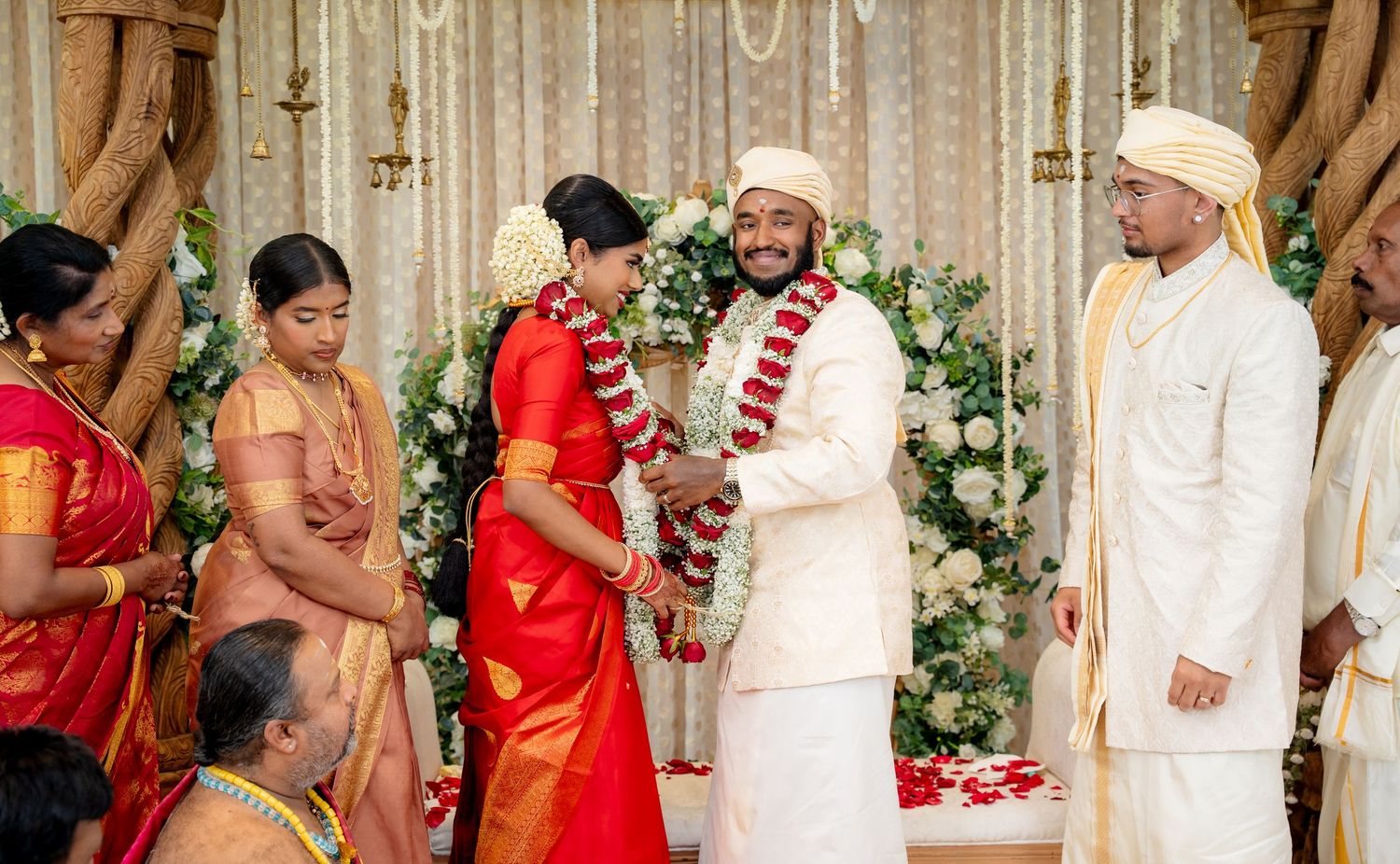 Smiling couple holds hands during vibrant South Indian Hindu wedding ceremony with floral decorations.
