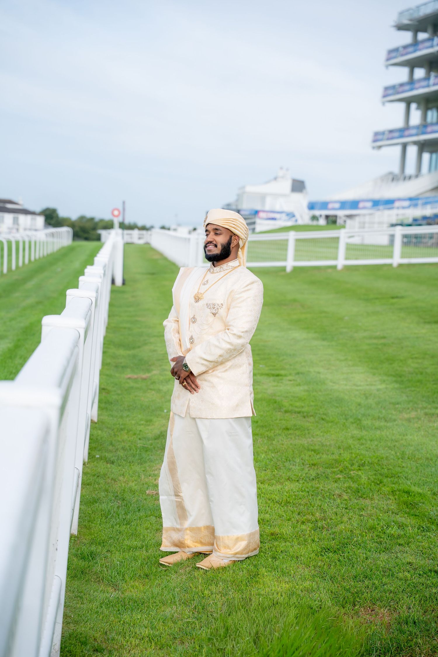 Smiling Indian groom in cream gold sherwani and turban standing on green grass at a racecourse.