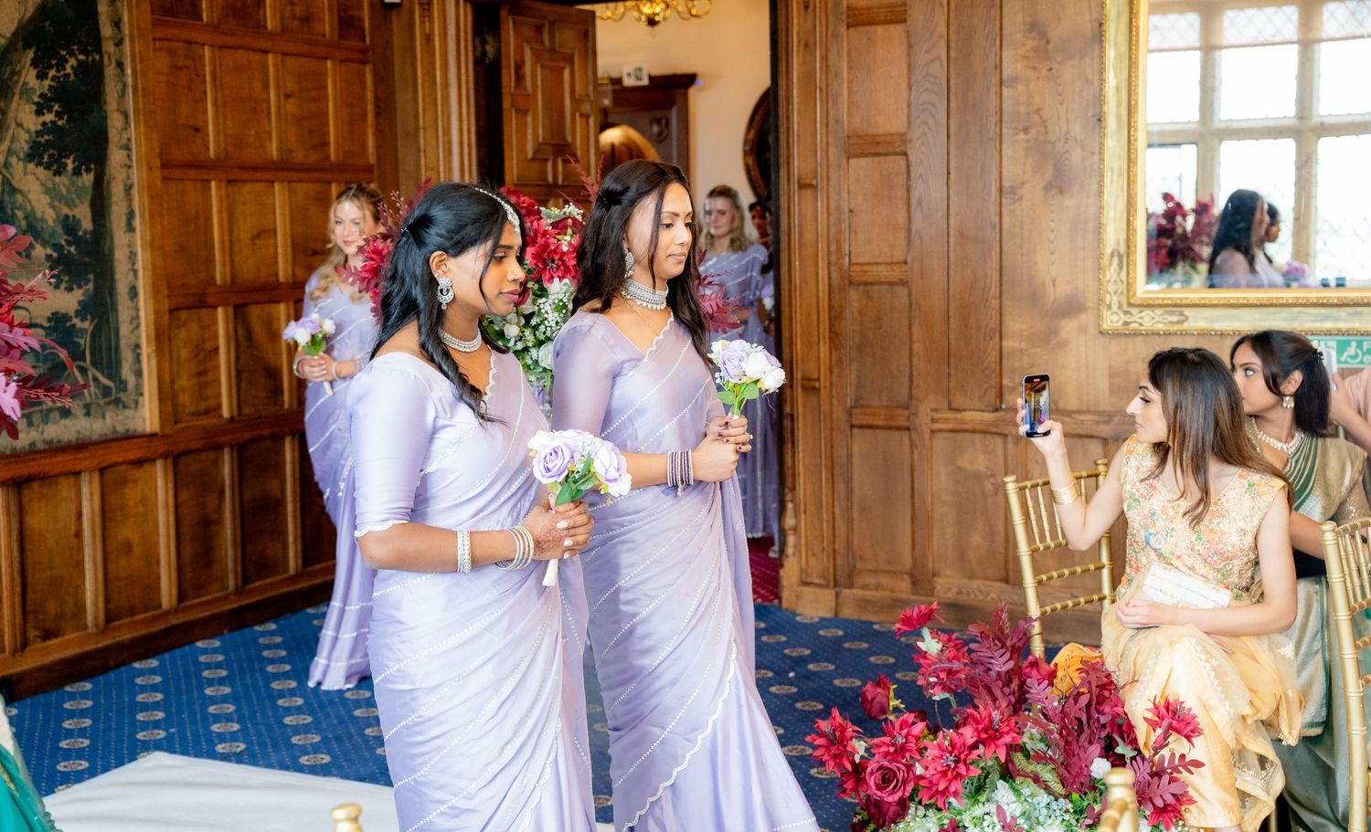 Two bridesmaids in lavender sarees holding white bouquets walk through an elegant wedding venue.