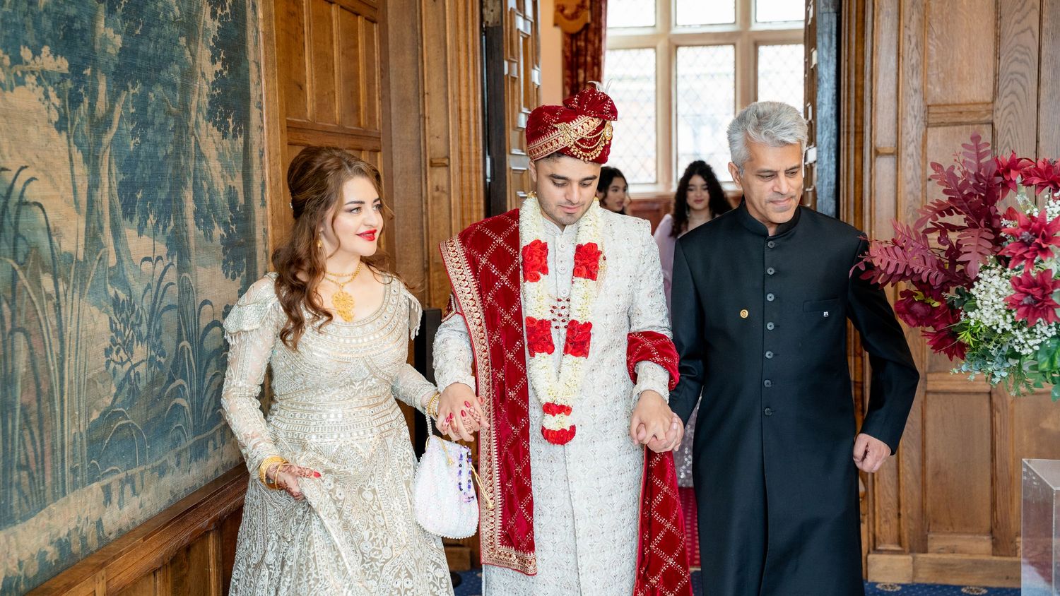 Groom in red sherwani walks with parents through decorated hall at Indian wedding ceremony.