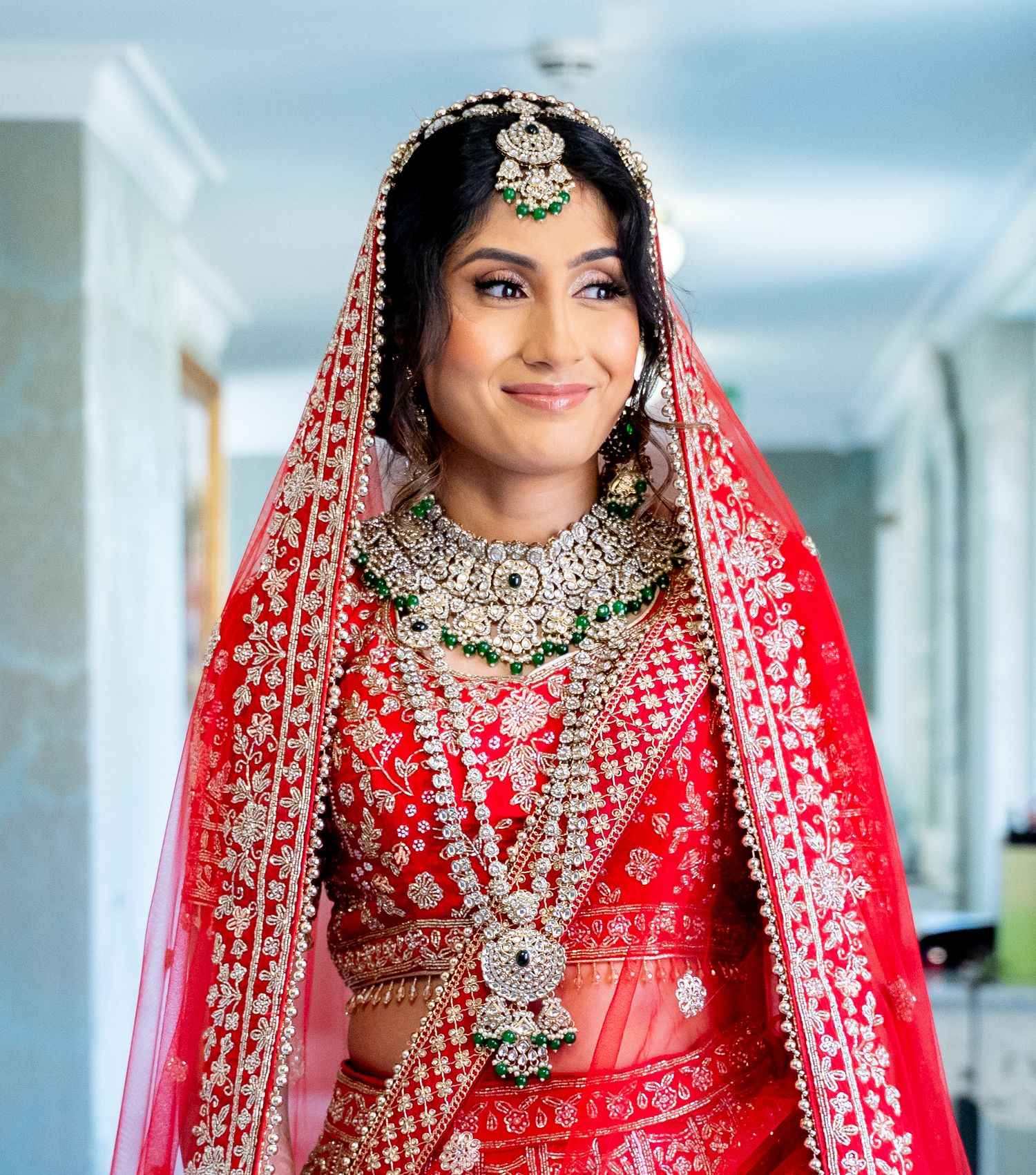 Indian bride smiling in red lehenga with green and gold bridal jewelry.