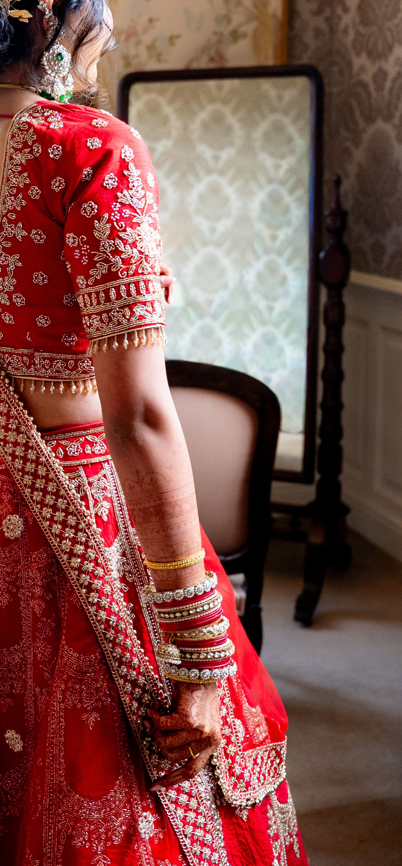 Indian bride in red embroidered lehenga with gold bangles stands before ornate mirror in elegant room.