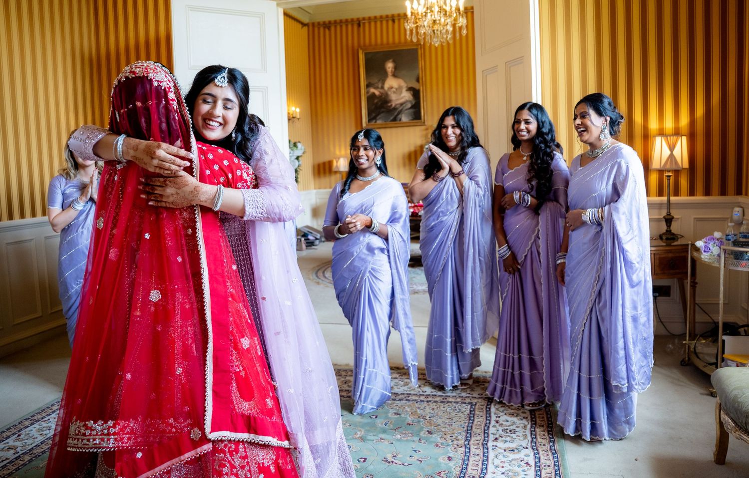 Indian bride embraced by bridesmaid while others in lavender sarees look on.