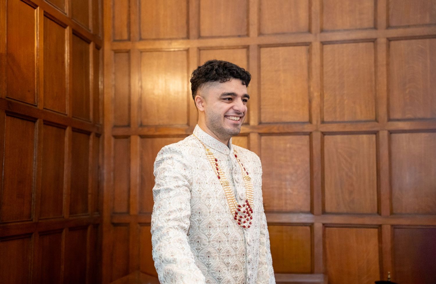 Handsome Indian groom smiling in traditional white embroidered sherwani against wooden paneled wall.