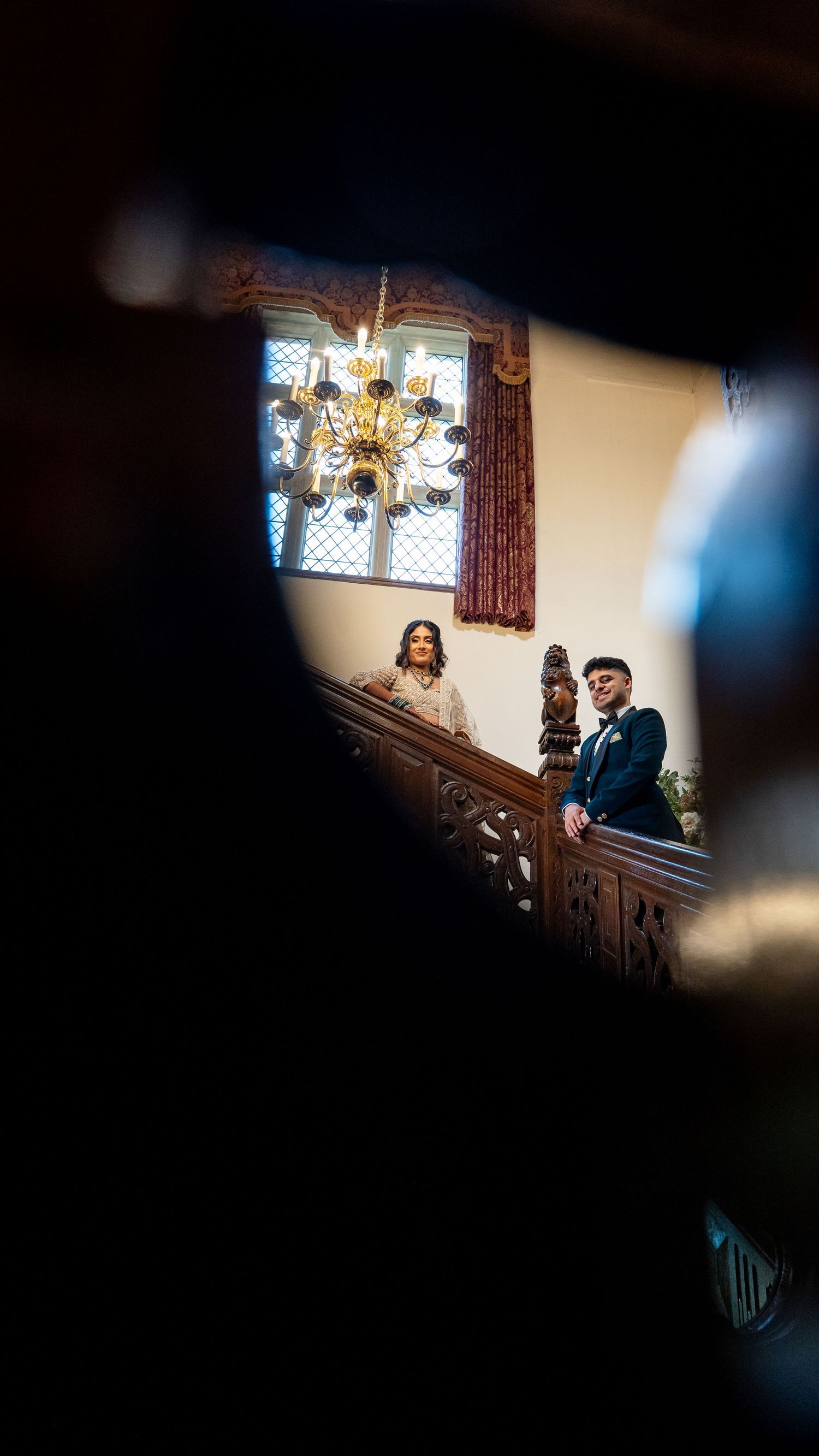 Bride and groom stand together at ornate wooden staircase balcony beneath golden chandelier at wedding.