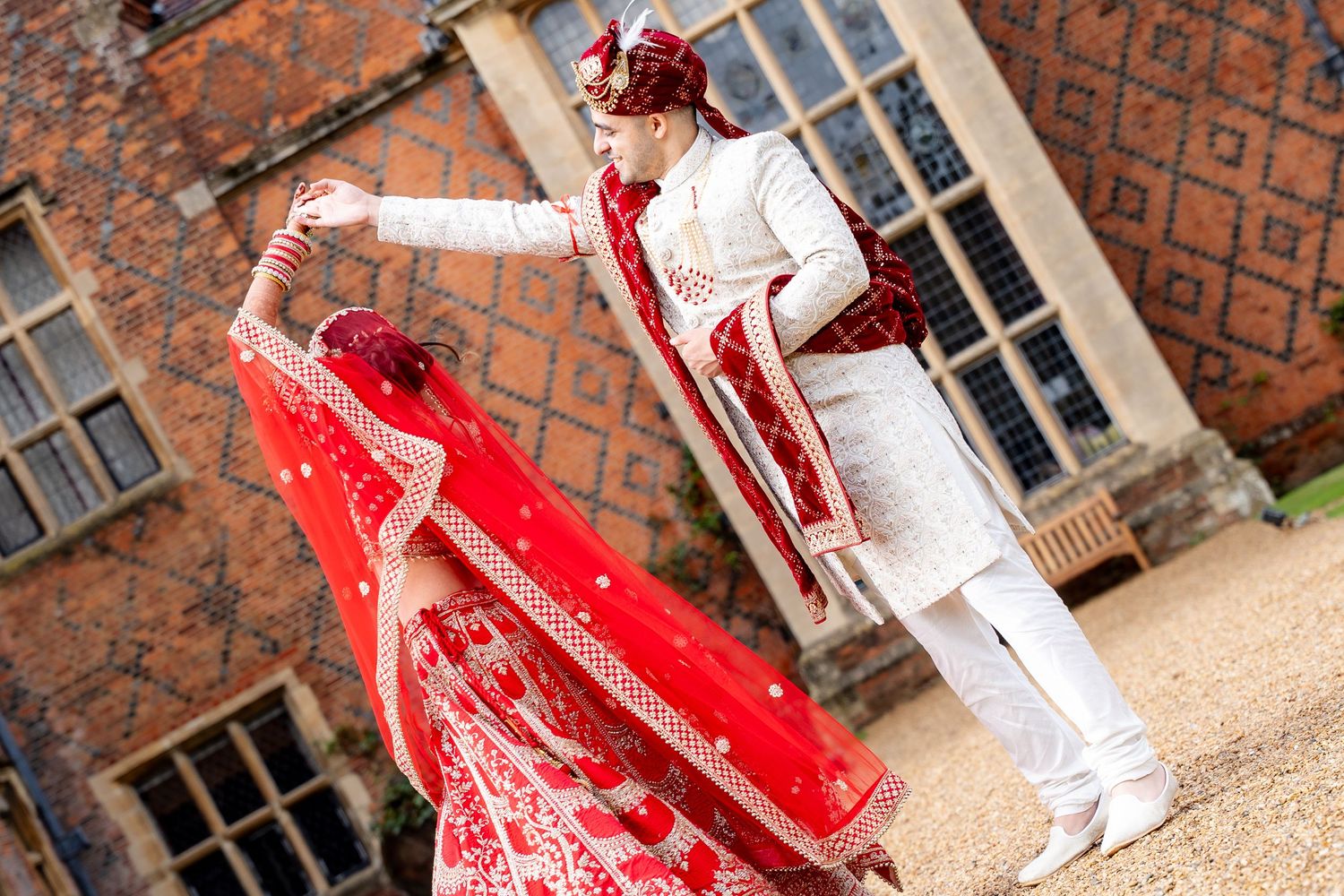 Indian bride in red lehenga twirling with groom in white sherwani outside historic brick venue.
