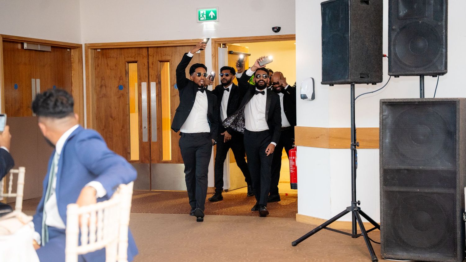 Groomsmen in black tuxedos make a grand entrance at a wedding reception hall with raised hands.