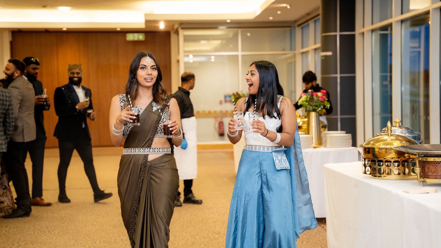 Two women in elegant Indian attire laughing and socializing at a formal event reception.