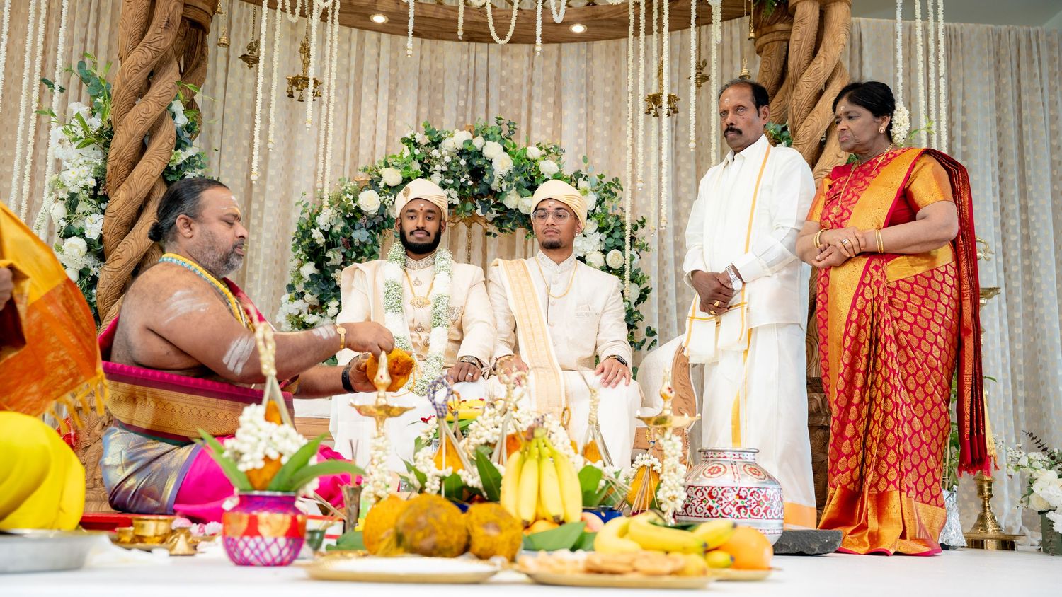Traditional Hindu wedding ceremony with groom in white sherwani at elegantly decorated mandap with white floral backdrop.