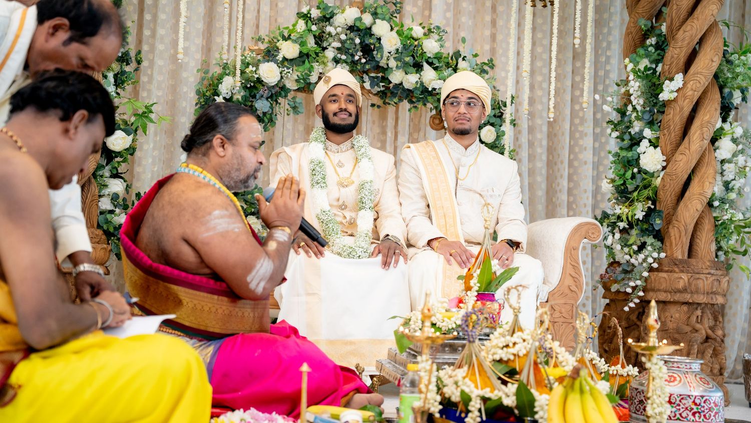 Two grooms smile during a Hindu wedding ceremony surrounded by priests and ritual offerings.