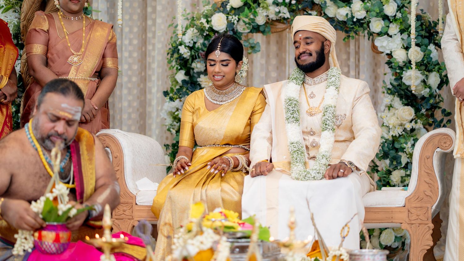 Tamil Hindu wedding couple in traditional attire seated during sacred ceremony rituals with priest officiating.