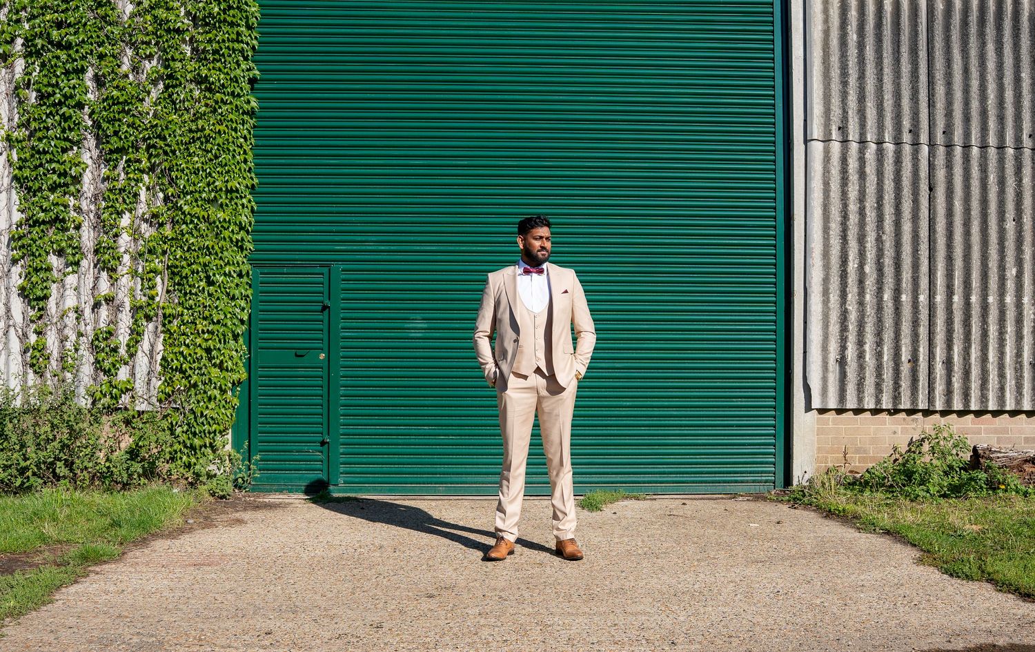 Groom in beige suit stands confidently before a green barn door in sunlight.