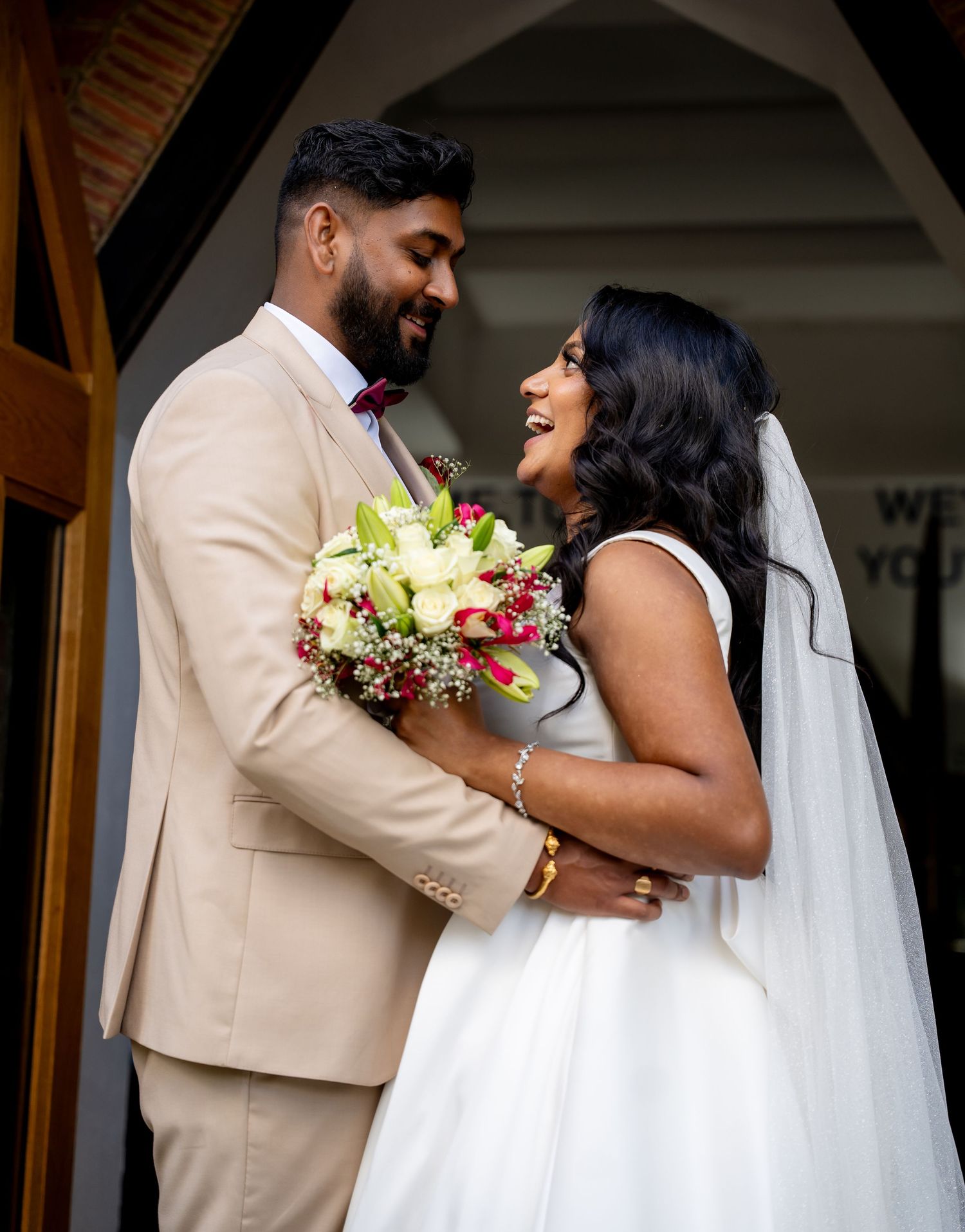 A joyful couple in wedding attire embraces, the bride holding a bouquet of white and red flowers, smiling at each other.