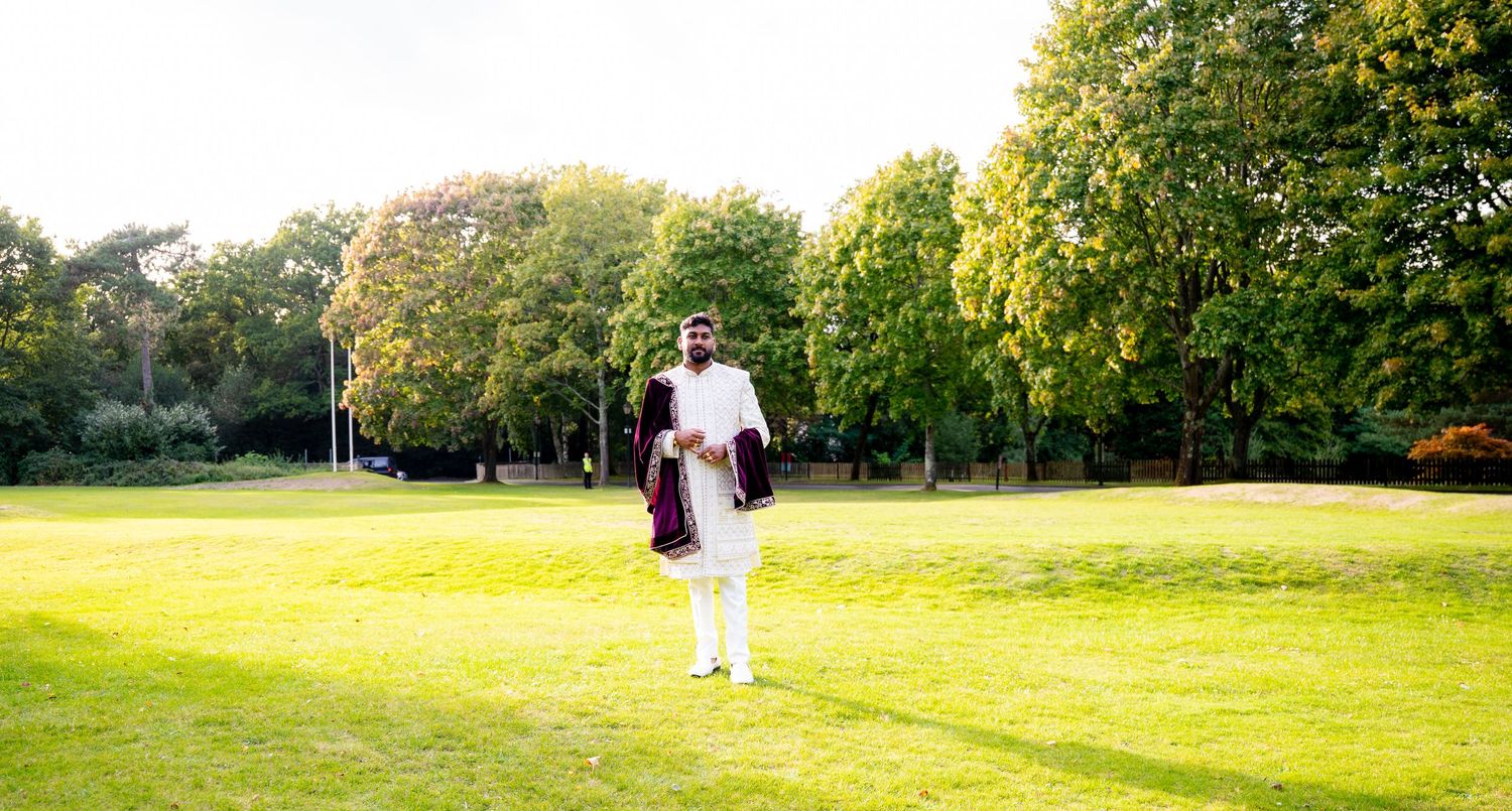 Groom in white sherwani with purple velvet shawl standing alone on green lawn surrounded by trees at Indian wedding.