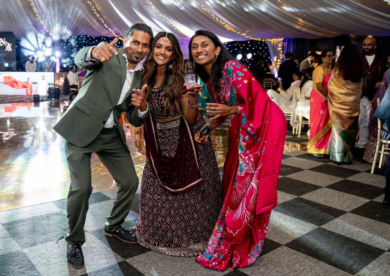 Three guests in Indian wedding attire posing and smiling on checkered dance floor at reception.