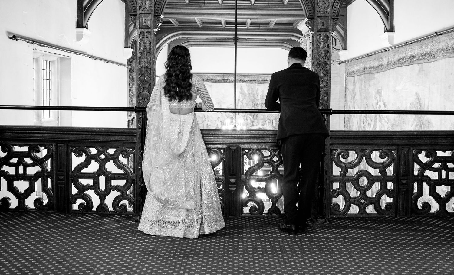 Black and white photo of a bride in a lehenga and groom in a suit standing at an ornate balcony railing.