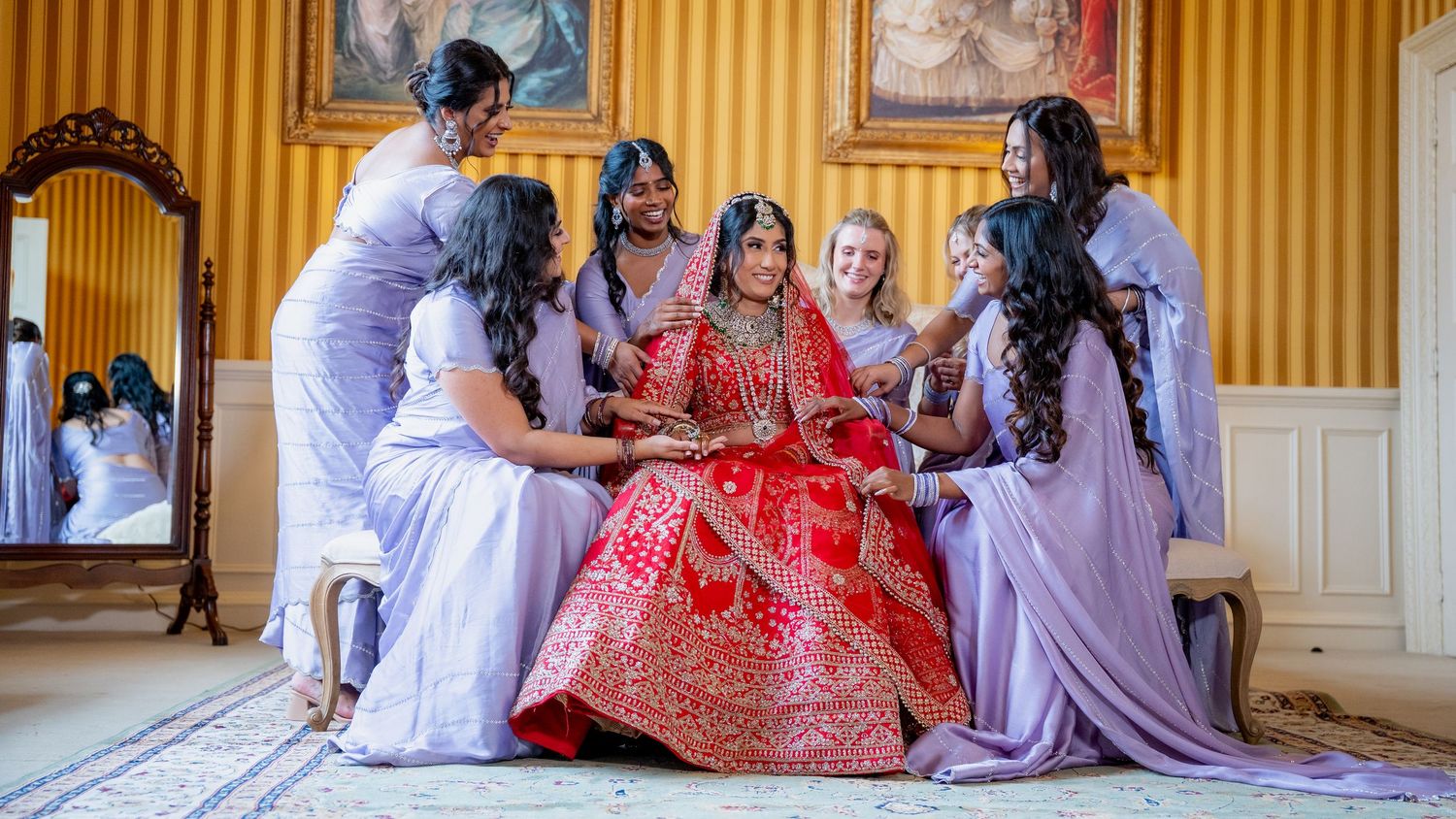 Indian bride in red lehenga sits surrounded by bridesmaids in lavender in golden room.