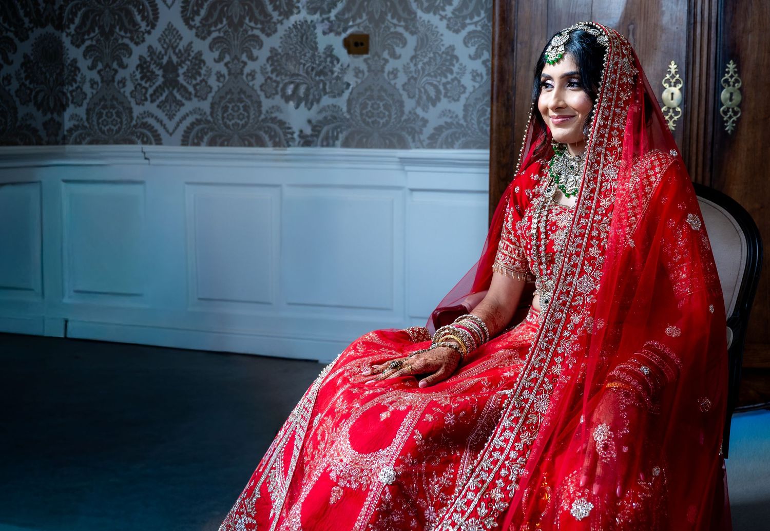 Indian bride in red bridal lehenga with green jewelry seated against decorative dark wallpaper.