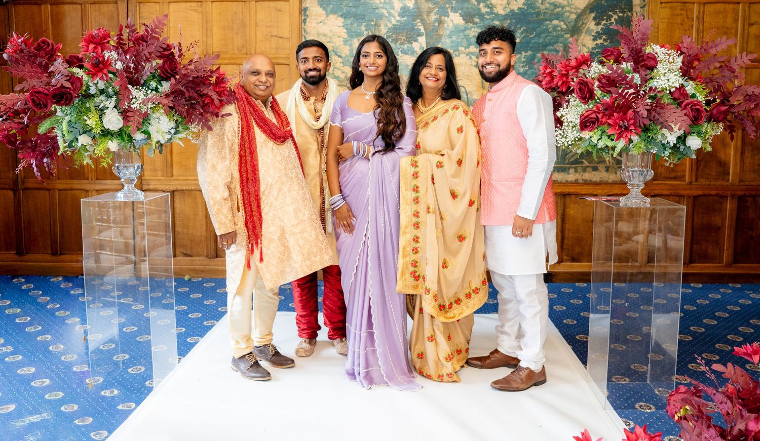 Family of five posing together at an Indian wedding ceremony with red floral arrangements.