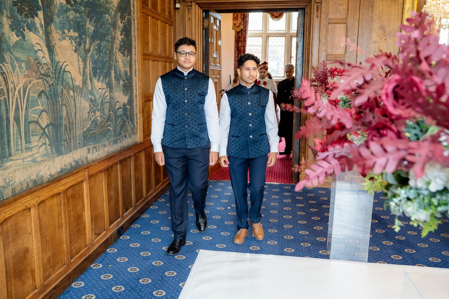 Two men in matching navy Nehru jackets walk through elegant carpeted corridor at Indian wedding.