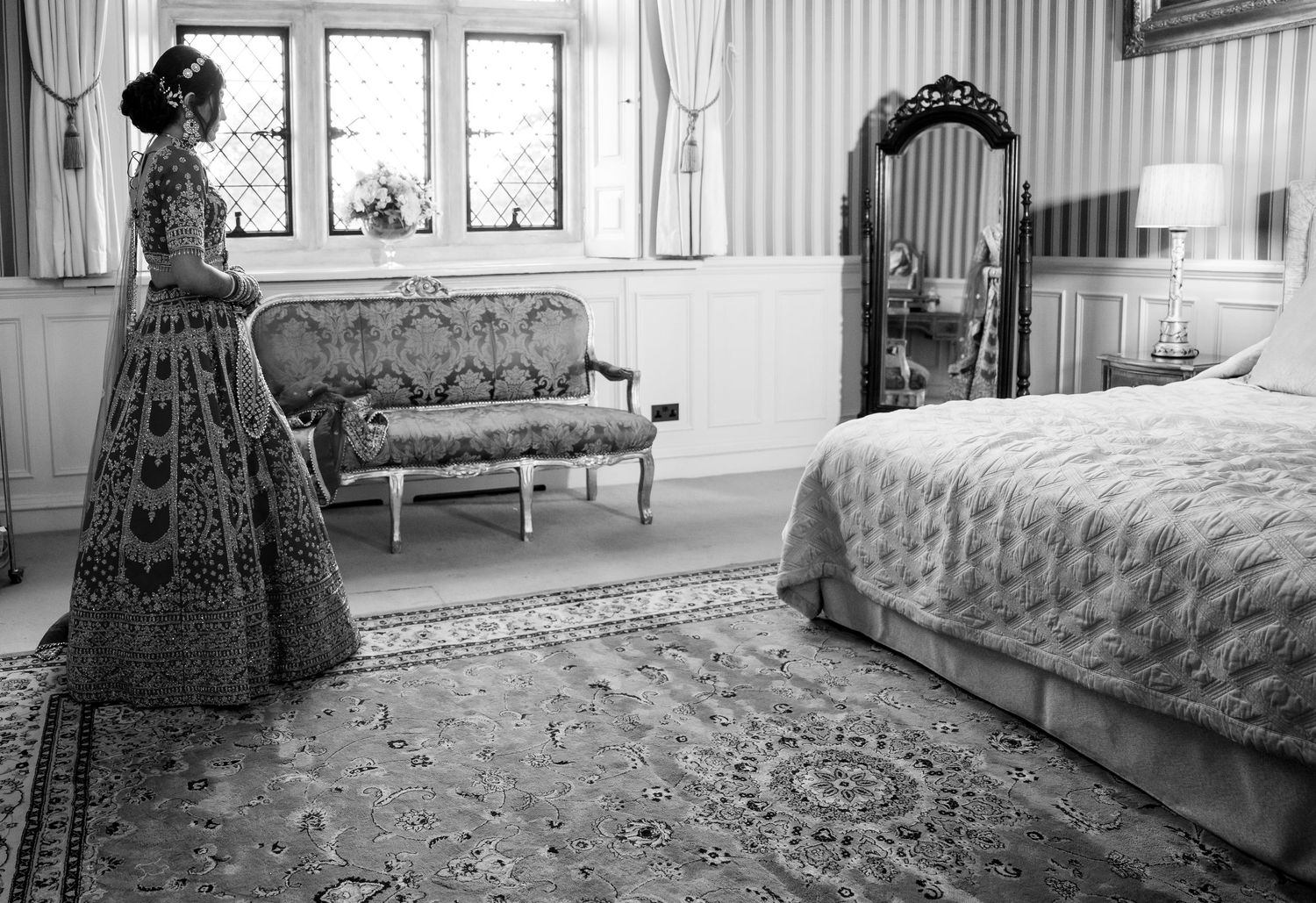 Woman in ornate gown stands in elegant bedroom with antique mirror and sofa, black and white photo.