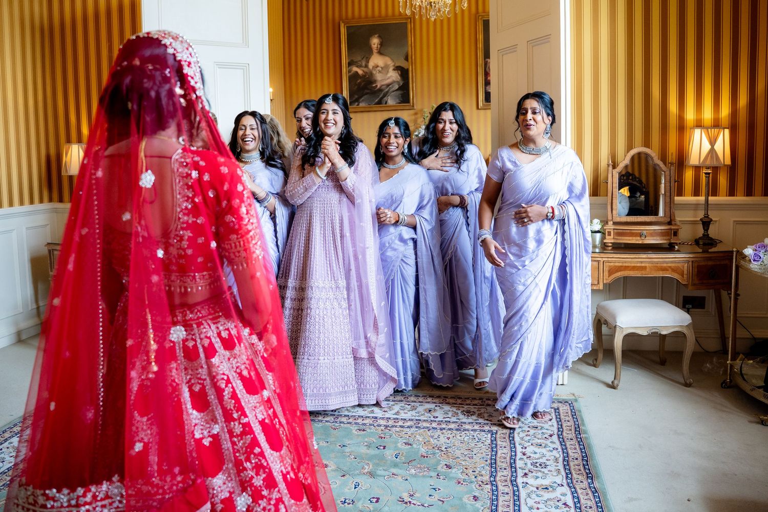 Indian bride in red lehenga with bridesmaids in lavender sarees reacting.