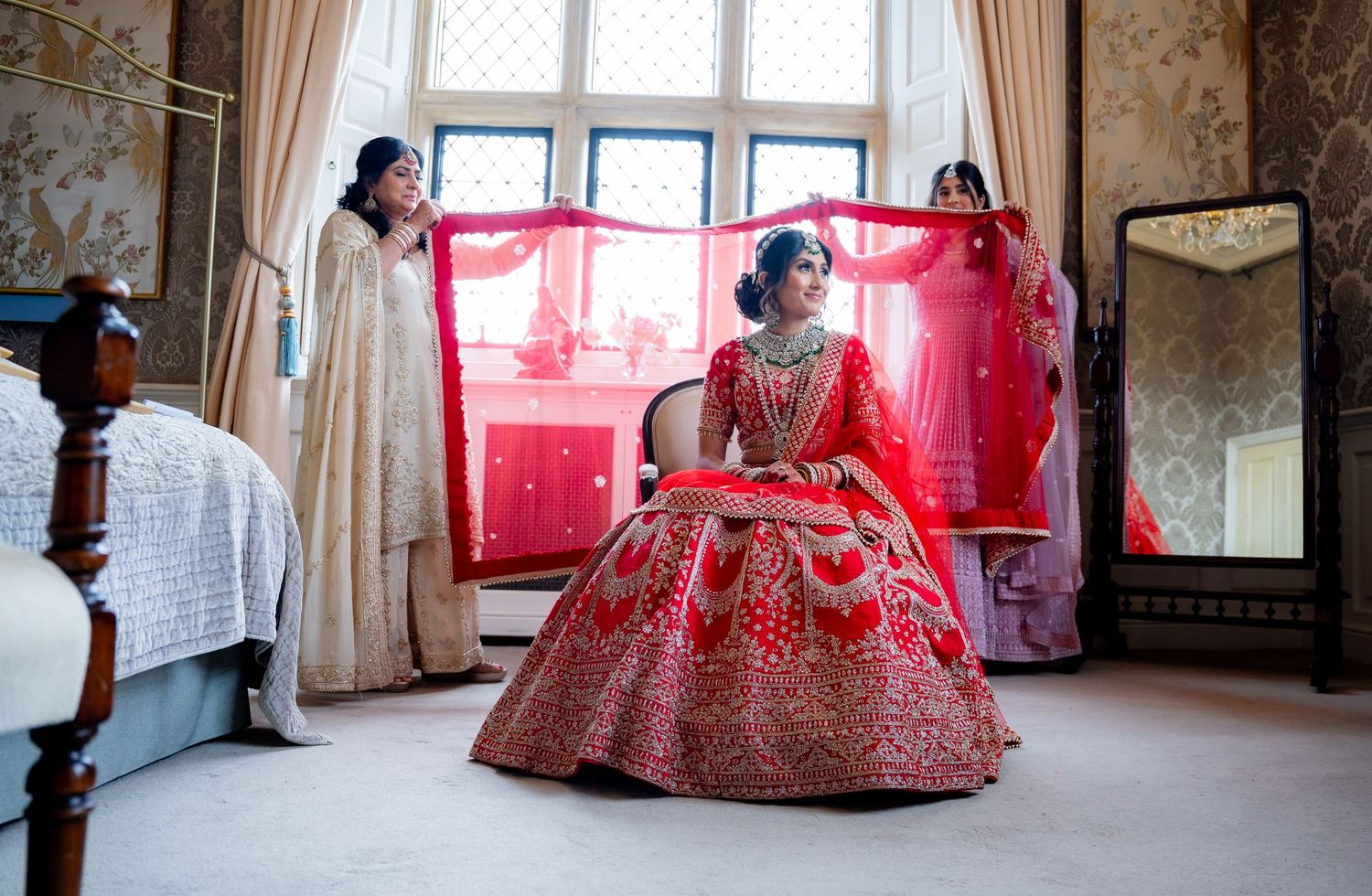 Indian bride in red lehenga seated as bridesmaid holds dupatta in elegant bedroom.