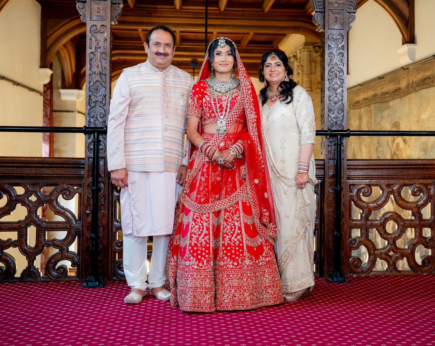 Indian bride in red lehenga posing with parents on decorative wooden balcony at wedding venue.